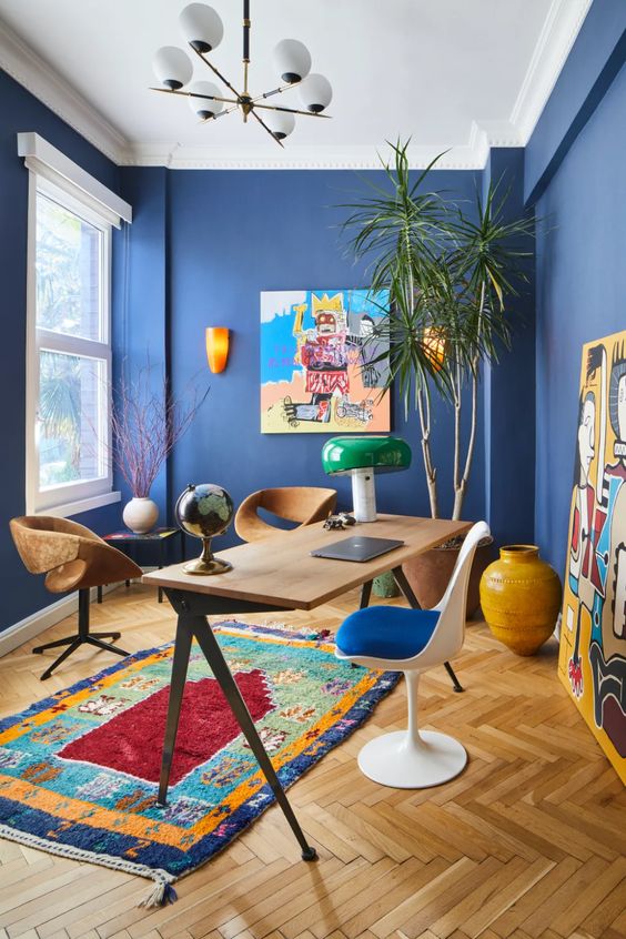 a vibrant home office with bold blue walls, colorful artworks and a rug, mismatching chairs and a statement plant in the corner