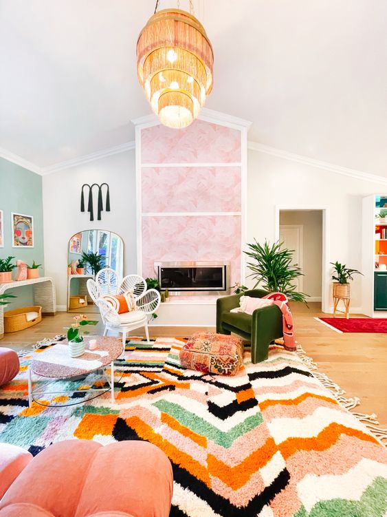 a vibrant maximalist living room with a pink clad fireplace, a green accent wall, a bold bold printed rug, coral furniture and a tiered fringe chandelier