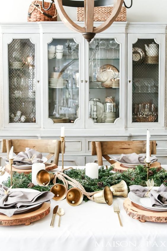 a vintage rustic Christmas tablescape with wooden slices, white porcelain, an evergreen, pinecone and bell garland and pillar candles