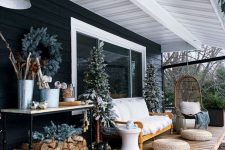 a winter terrace with a stained wooden deck, a shelf for storage, a sofa, jute poufs, a hanging egg-shaped chair, a couple of trees and an evergreen wreath