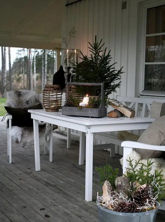 a winter terrace with white furniture, faux fur blankets and neutral pillows, candles, pinecones, evergreens and candle lanterns is super welcoming