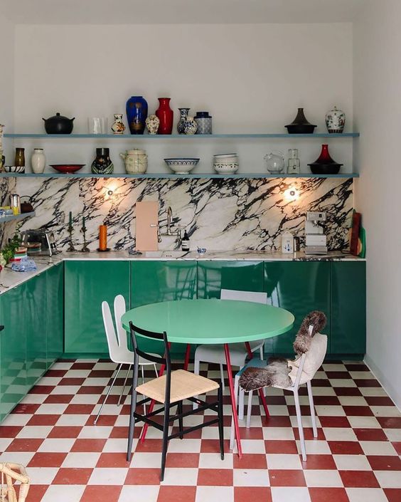 an elegant maximalist kitchen with emerald cabinets, a red and white checked floor, a white marble backsplash and countertop, floating shelves with various decor