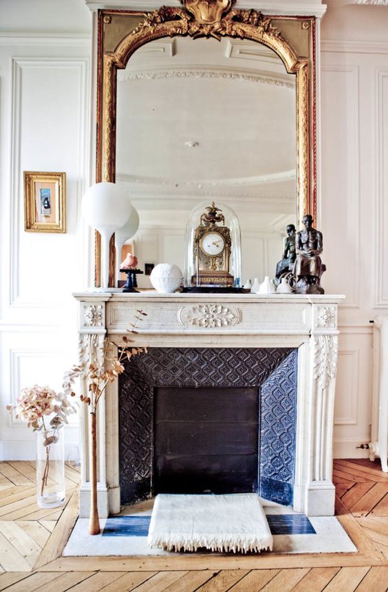 a fabulous vintage fireplace with an ornated white mantel, an oversized mirror and some vintage decor on the mantel