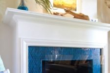 06 a fireplace surrounded with bright blue tiles, with an elegant white mantel and some coastal art on it