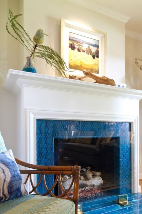 a fireplace surrounded with bright blue tiles, with an elegant white mantel and some coastal art on it