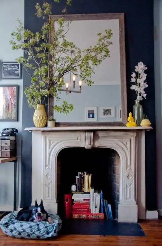 a navy wall with a brick fireplace and an ornated white wooden mantel that accents it, colorful books inside and bold yellow vases help it to stand out