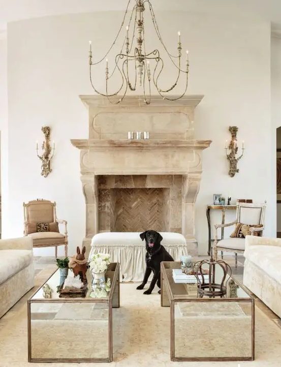 a vintage-inspired French chic living room with neutral furniture, a large stone and brick clad fireplace, a large chandelier and modern mirror tables