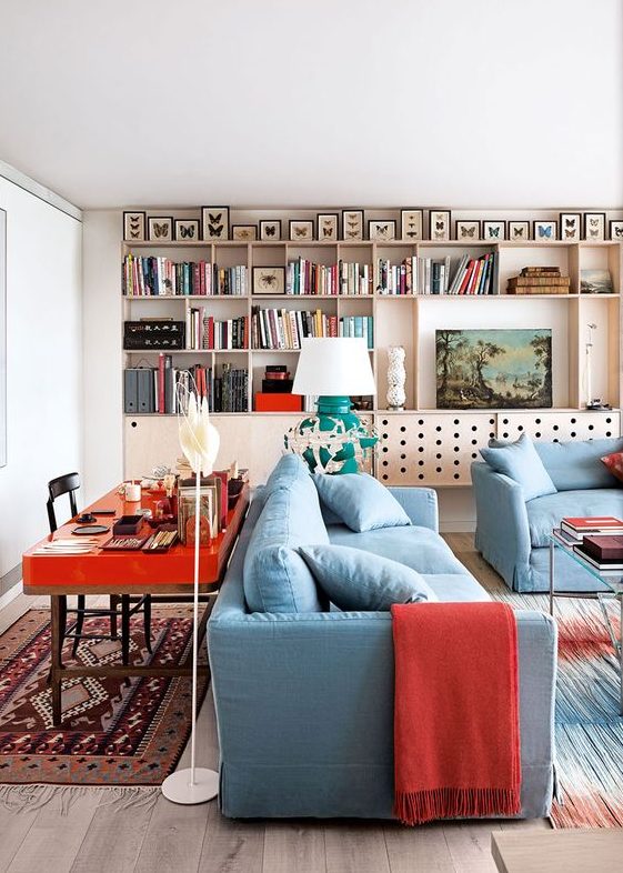 a bold living room with a plywood storage unit on the wall, butterflies, blue furniture, a hot red desk behind the sofa and bold red textiles