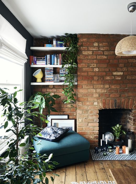 a modern living room with a red brick accent wall with a fireplace, niche shelves, potted greenery, a cozy blue ottoman