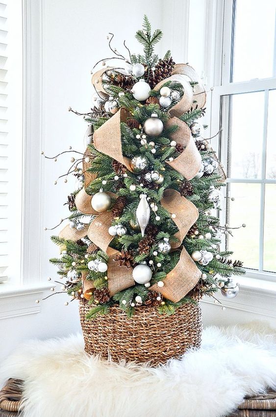 a lovely tabletop woodland rustic Christmas tree with silver and white ornaments, pompoms, burlap ribbon, pinecones placed into a basket