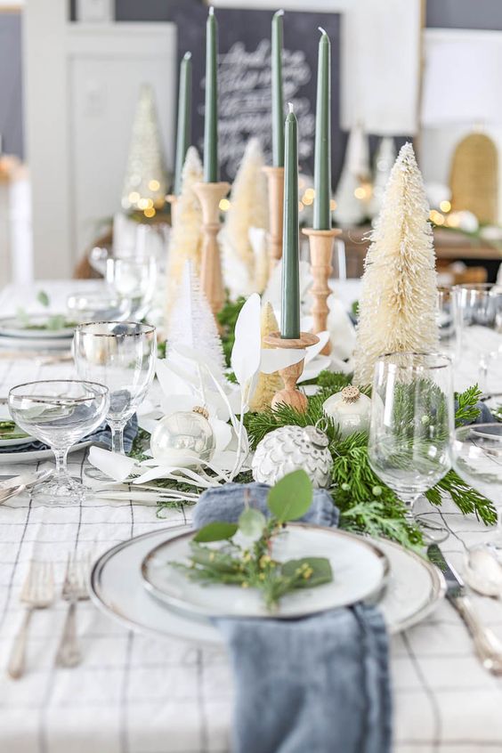 a neutral Christmas tablescape with a plaid tablecloth and blue napkins, neutral bottle brush trees, green candles and simple porcelain