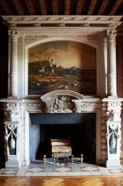 a gorgeous ornated antique fireplace with pillars and a painting surrounding it and some tiles on the floor in front of it is really a royal feature