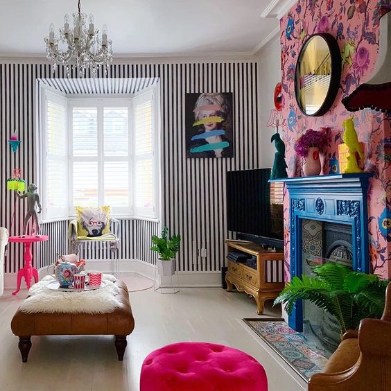 a maximalist living room with a striped wall, a bold floral one, a bright blue mantel over a vintage fireplace, a crystal chandelier and touches of hot pink