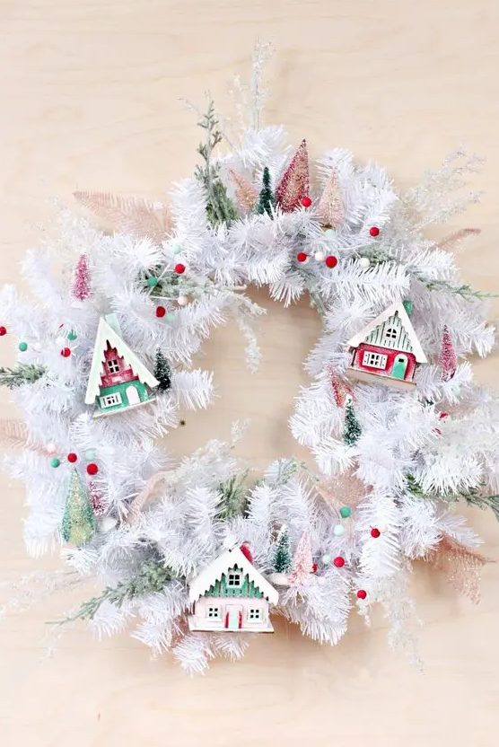 a white evergreen Christmas wreath with colorful pompoms, colorful small houses and bottle brush Christmas trees