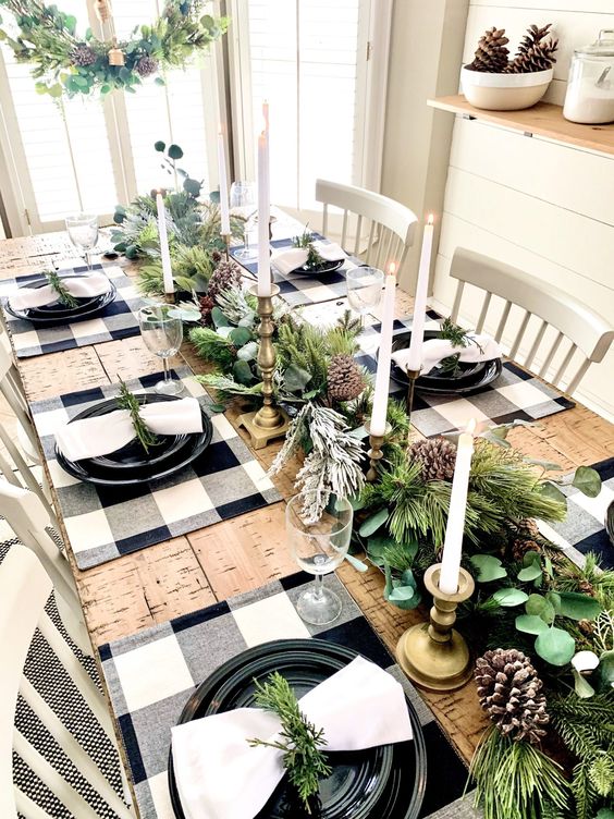 a natural Christmas table setting with buffalo check placemats, black plates, an evergreen garland with pinecones and candles
