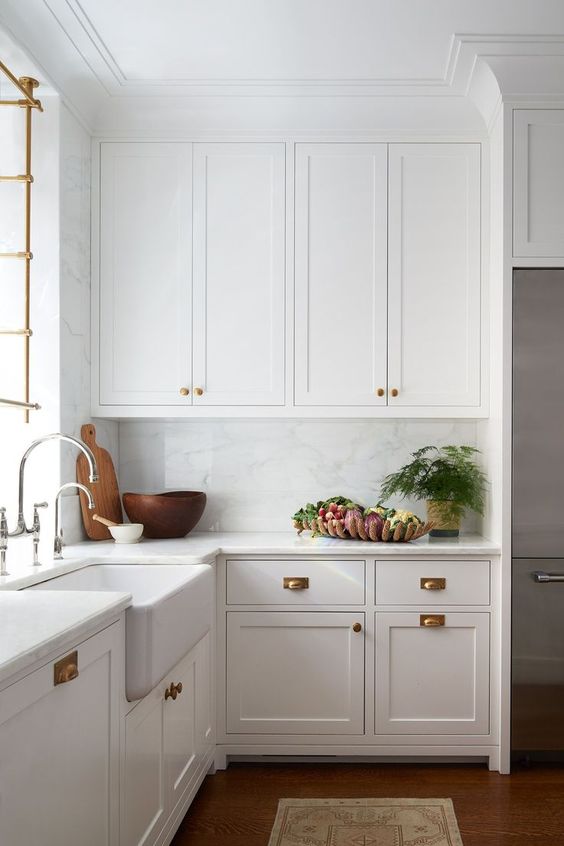 a beautiful white farmhouse kitchen with shaker cabinets, brass handles, crown molding that connects the cabinets with the ceiling and makes the space look higher
