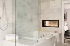 a beautiful white marble bathroom with large scale tiles covering the bathtub, too, a double-sided fireplace and neutral textiles