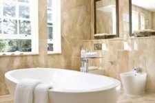 a beige bathroom fully clad with marble, with an oval tub, large mirrors and a window for more light