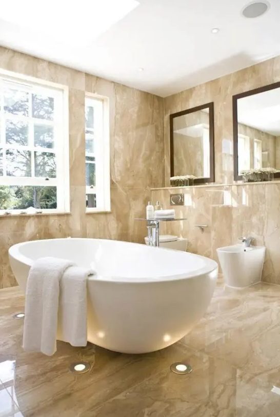a beige bathroom fully clad with marble, with an oval tub, large mirrors and a window for more light