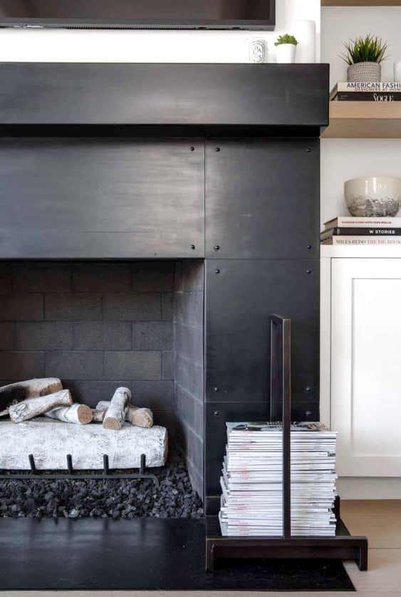 a brick fireplace surrounded with blackened metal sheets and with a magazine stand by its side is cool