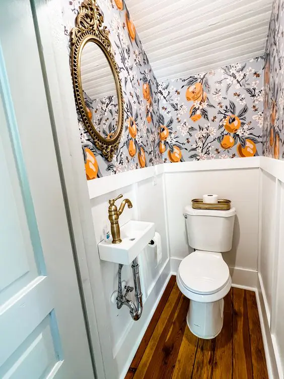 a chic and tiny under the stairs powder room with bold wallpaper and paneling, a wall-mounted sink and a mirror in a vintage frame