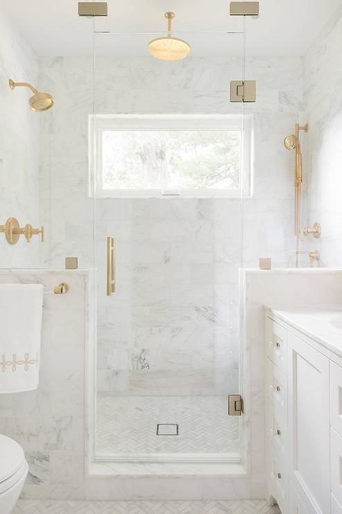 a chic white marble bathroom with herringbone and subway tiles, elegant brass fixtures and a small window in the shower