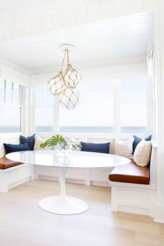 a coastal dining space with a view, a banquette seating with pillows, an oval table and buoy-style pendant lamps