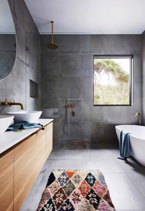 a contemporary bathroom clad with concrete tiles completely, a stained floating vanity, an oval tub, gold fixtures and a bold printed rug