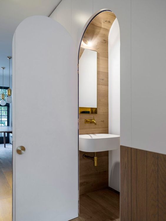 a contemporary powder room hidden under the stairs, with an arched door, a wall-mounted sink and some timber cladding the space