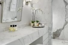 a contemproary luxurious bathroom done with white marble everywhere, with a sleek vanity and a large mirror