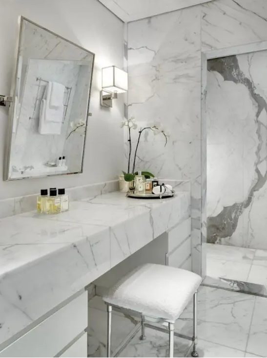 a contemproary luxurious bathroom done with white marble everywhere, with a sleek vanity and a large mirror
