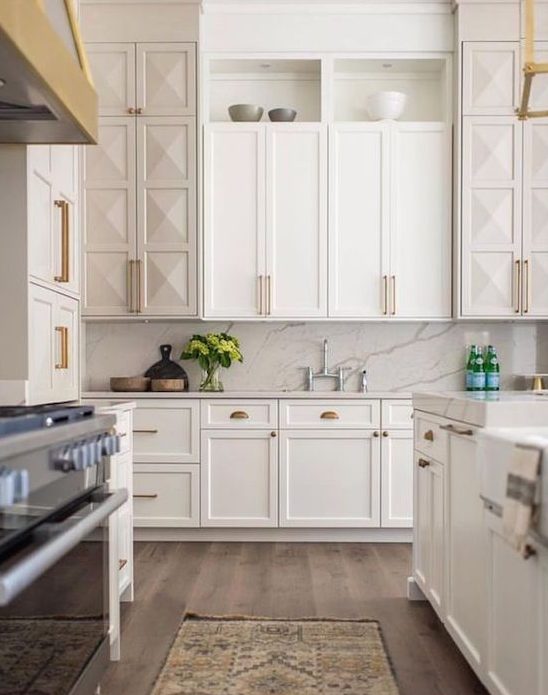 a creamy kitchen with shaker style and geometric cabinetry, a white marble backsplash and countertops plus gold and brass touches