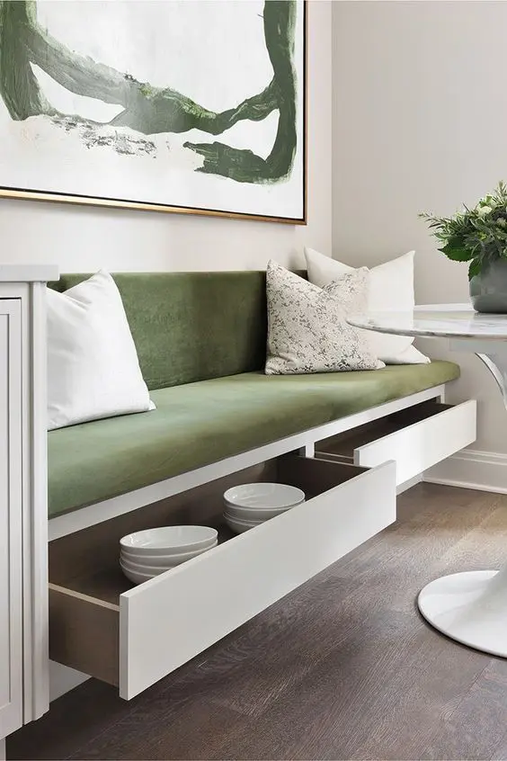 a cute dining nook with a banquette seating with storage drawers upholstered with green velvet, a round table and a bold artwork