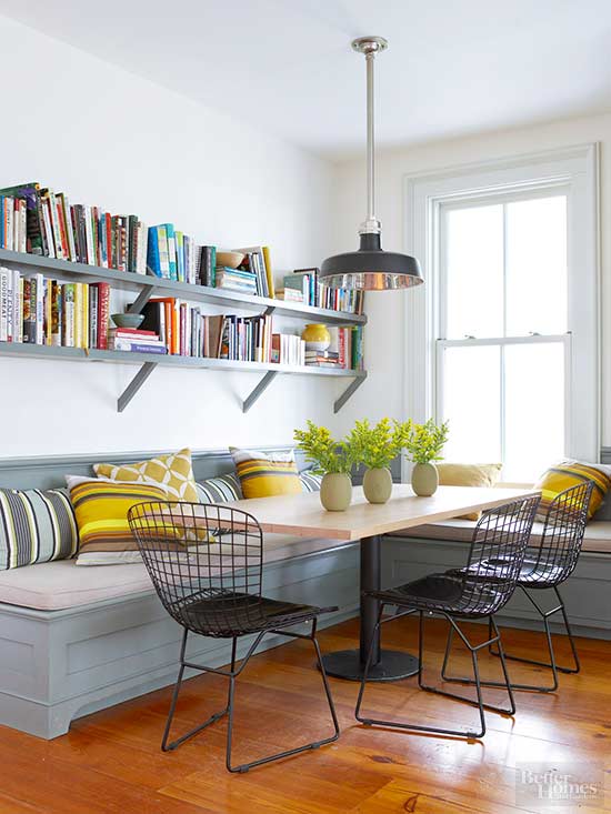 a farmhouse dining space with a grey banquette with colorful pillows, a table on a metal leg, open shelving, black metal chairs