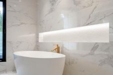 a jaw-dropping white marble bathroom with a lit up niche for storage, an oval tub, a gold faucet and built-in lights