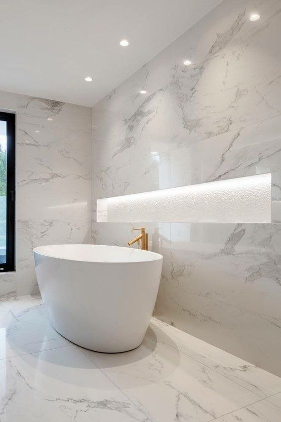 a jaw dropping white marble bathroom with a lit up niche for storage, an oval tub, a gold faucet and built in lights