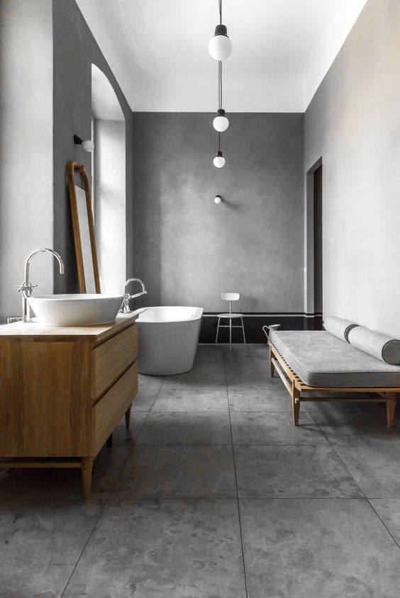 a large concrete bathroom with an elegant bathtub, a light-stained vanity with a round sink, a chic daybed and pendant lamps