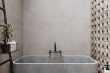 a lovely bathroom of concrete, with a terrazzo floor, a catchy accent wall and a concrete bathtub plus a black ladder