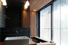a minimalist bathroom done with light stained wooden slabs and black skinny tiles, with pendant lamps and a large window for more light