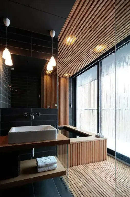 a minimalist bathroom done with light stained wooden slabs and black skinny tiles, with pendant lamps and a large window for more light