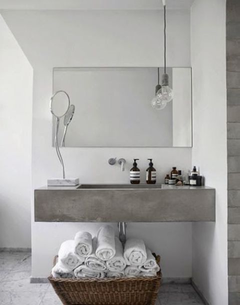 a minimalist bathroom with white plaster walls, a concrete floating sink, a basket with towels and a laconic mirror