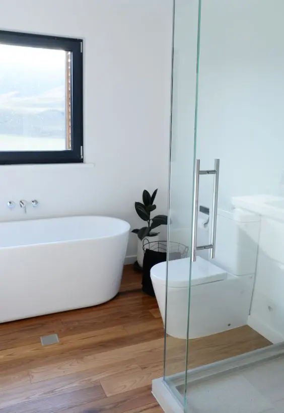a minimalist white bathroom with a light-stained wood floor, a shower space, an oval tub and some dark touches for more drama
