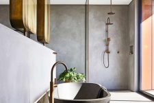 a modern bathroom done with concrete walls, floor, an oval tub, brass fixtures and a glazed wall for much light