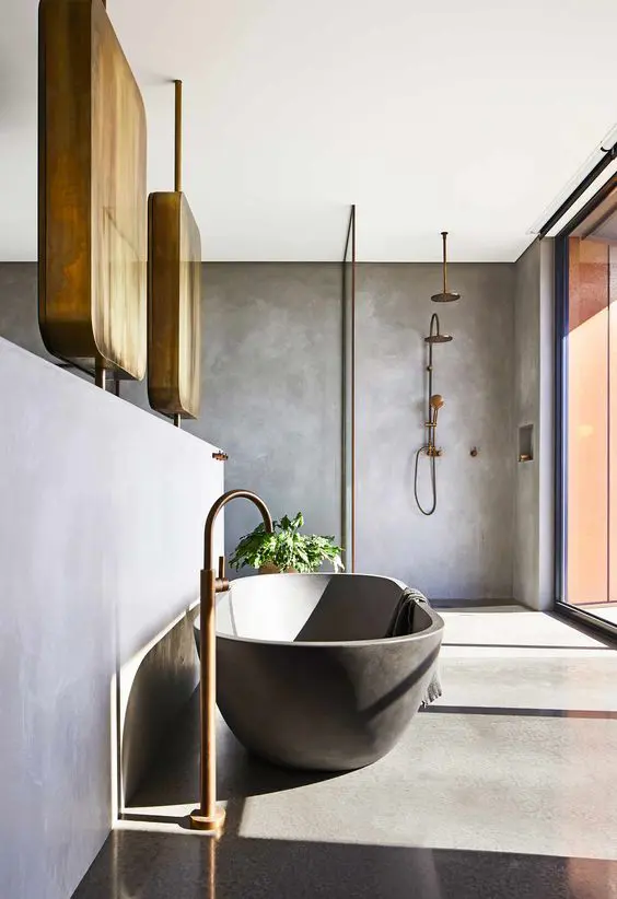 a modern bathroom done with concrete walls, floor, an oval tub, brass fixtures and a glazed wall for much light