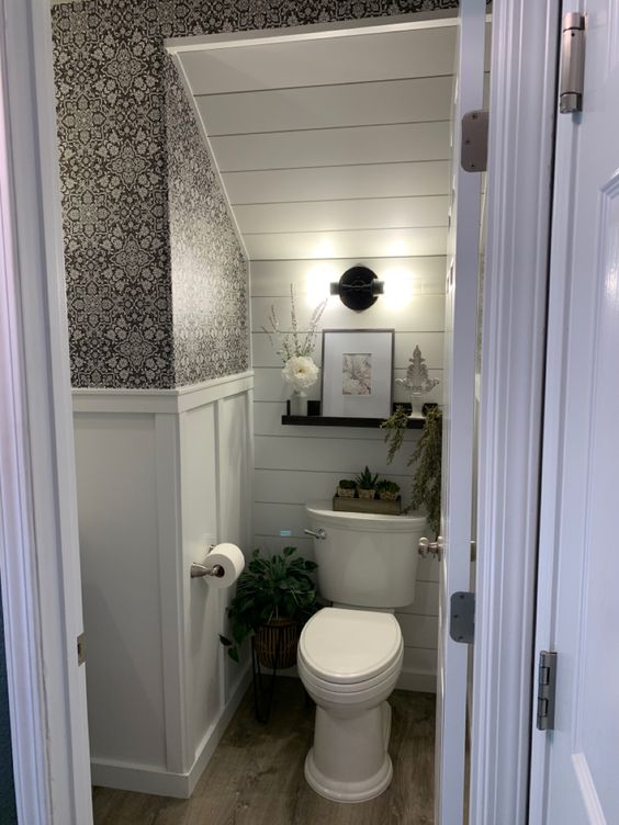 a modern farmhouse powder room under the stairs, with a shiplap wall, printed wallpaper, an open shelf and some decor and greenery
