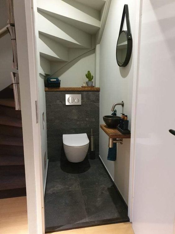 a modern powder room under the stairs clad with grey tile, a wall-mounted toilet, a black bowl sink and a mirror