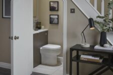 a modern powder room with greige walls, a taupe vanity, a toilet and some artwork under the staircase