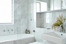 a neutral contemporary bathroom clad with marble tiles, with a tub, a toilet and a large mirror cabinet on the wall