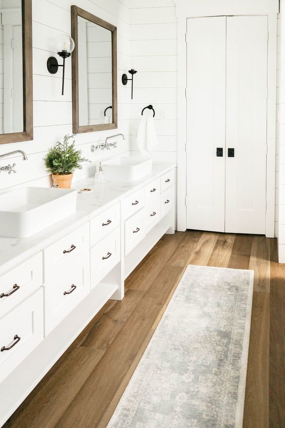 a neutral modern farmhouse bathroom with a light-stained wooden floor, a large double vanity, mirrors in reclaimed wood frames and vintage sconces