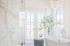 a neutral sophisticated bathroom with a dose of luxury – a white marble shower space, a parquet floor, an oval tub, a crystal chandelier and windows and a door to a terrace